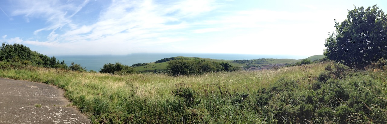 Uitzicht over de zee vanaf de kliffen van Dover. Het landschap is glooiend en begroeid met ruig gras. De lucht is lichtbewolkt. In de verte zie je de kust van Frankrijk.