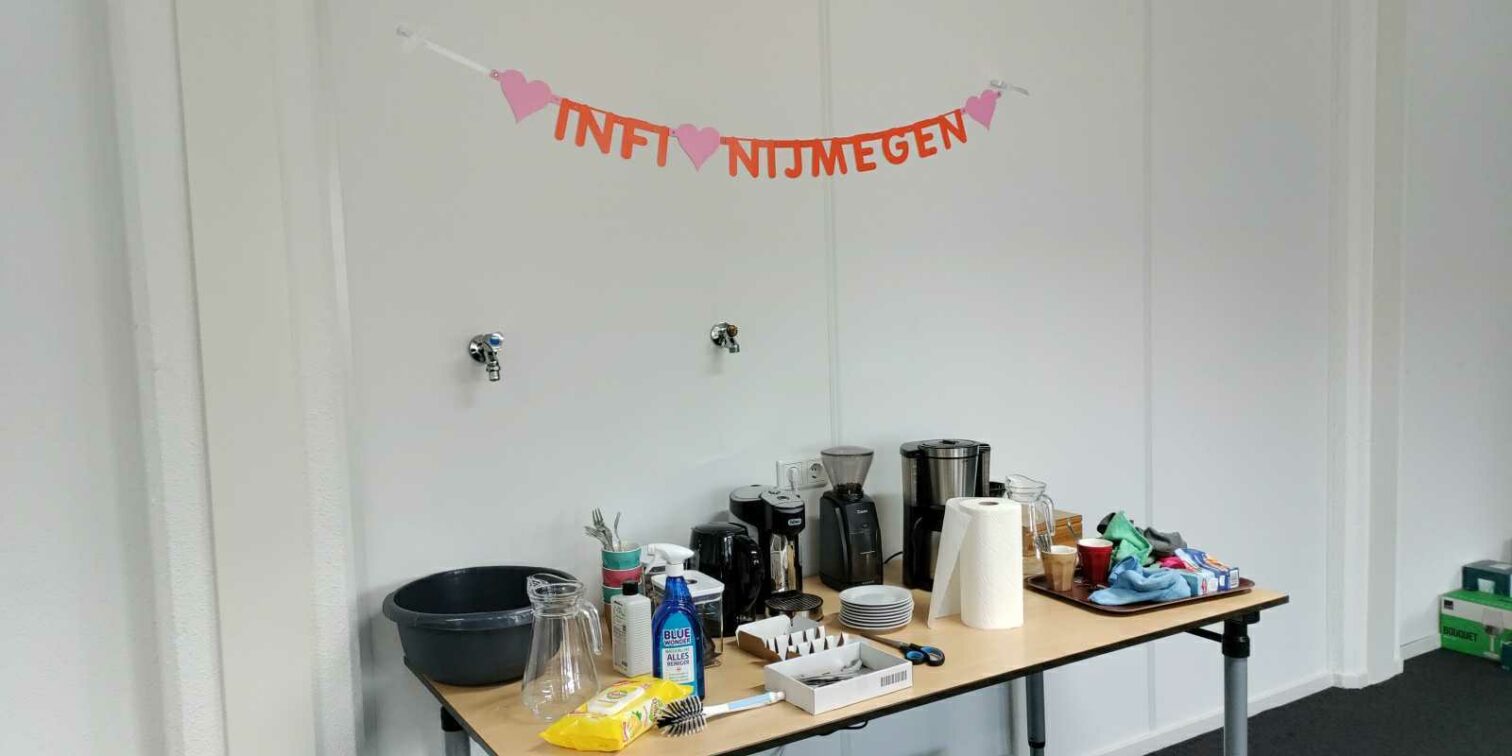 Het kantoor van Nijmegen, met een volle tafel met keukenspullen, koffiezetapparaat, etc. Aan de muur hangt een slinger met de tekst "Infi Nijmegen"."