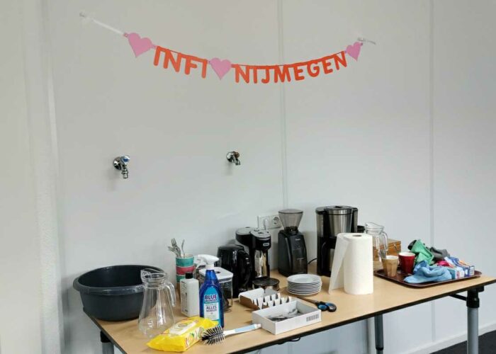 Het kantoor van Nijmegen, met een volle tafel met keukenspullen, koffiezetapparaat, etc. Aan de muur hangt een slinger met de tekst "Infi Nijmegen"."