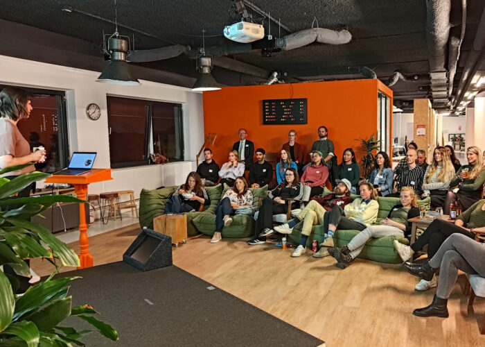 De spreker Lisanne van der Palen en het publiek tijdens de meetup. De spreker staat links op het podium en kijkt naar het publiek. Het publiek op de foto bestaat uit ongeveer 30 mensen, meer vrouwen dan mannen. Ze zitten op stoelen en banken en hebben allemaal een naamkaartje op. Achter het publiek hangt een board bestaande uit gekleurde grote pixels, die het woord 'UX!' spellen.