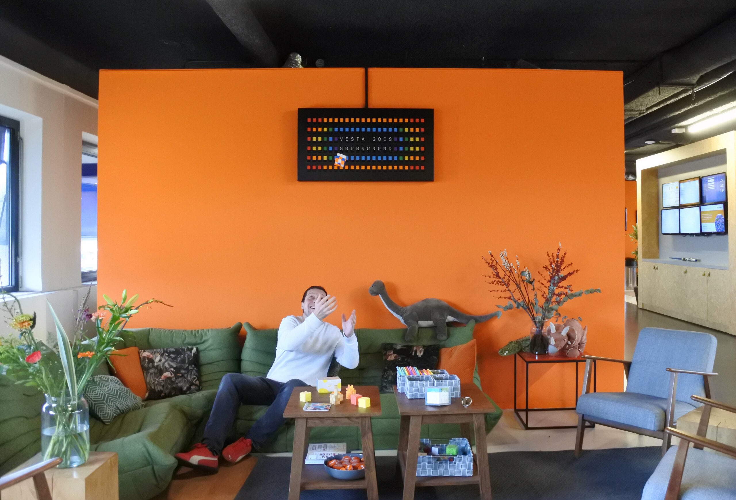 Oranje muur met bovenaan een Vestaboard met kleurrijk beeld, op de groene bank ervoor een lachende man die een gegooide rubikscubus probeert op te vangen.