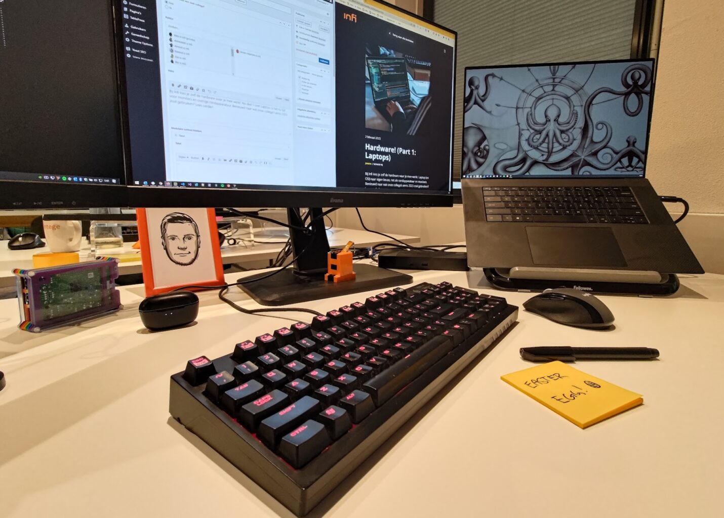 Werkplek met twee monitors, een laptop op een standaard, en vooraan prominent een toetsenbord en muis. Plus een post-it waar "Easter Egg!" op geschreven staat in het hoekje.