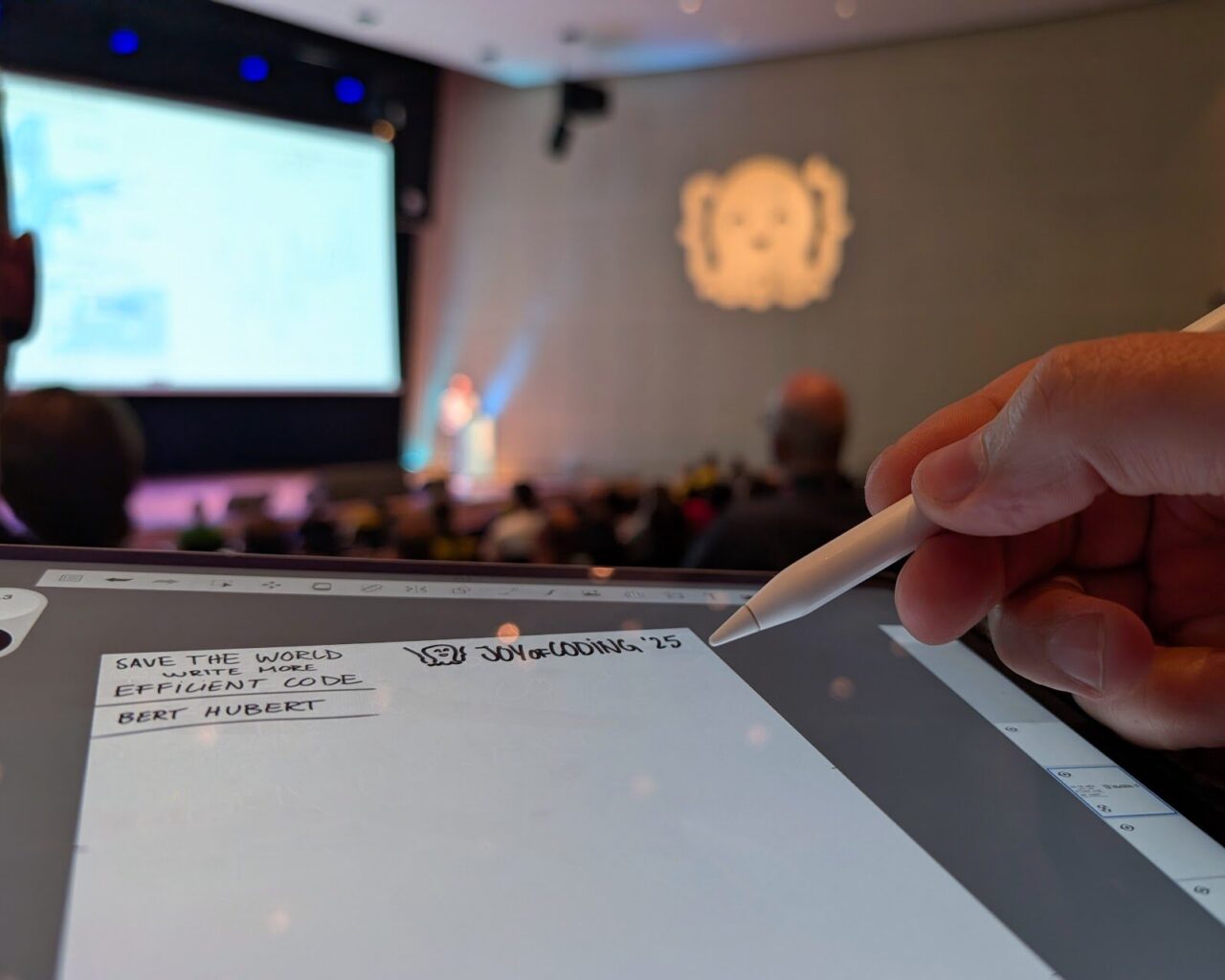 Close up van een tablet waarop getekend wordt met witte pen, conferentiezaal op de achtergrond met blurry Octopus-icoon op de muur geprojecteerd
