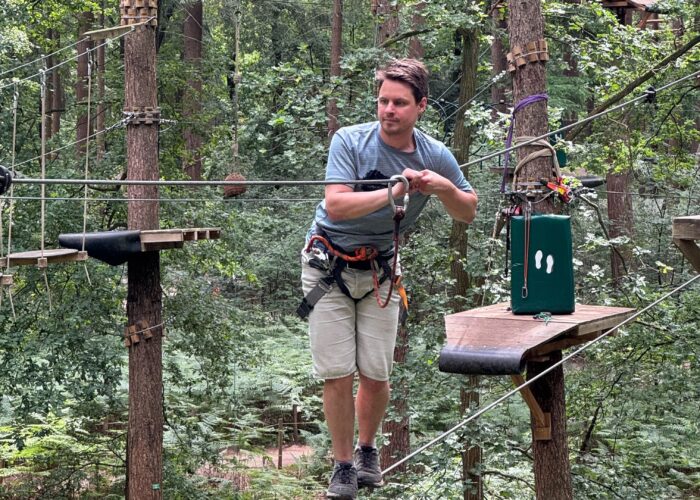 Persoon die geconcentreerd kijkt terwijl hij in klim-outfit over twee touwen balanceert. Bos op de achtergrond.