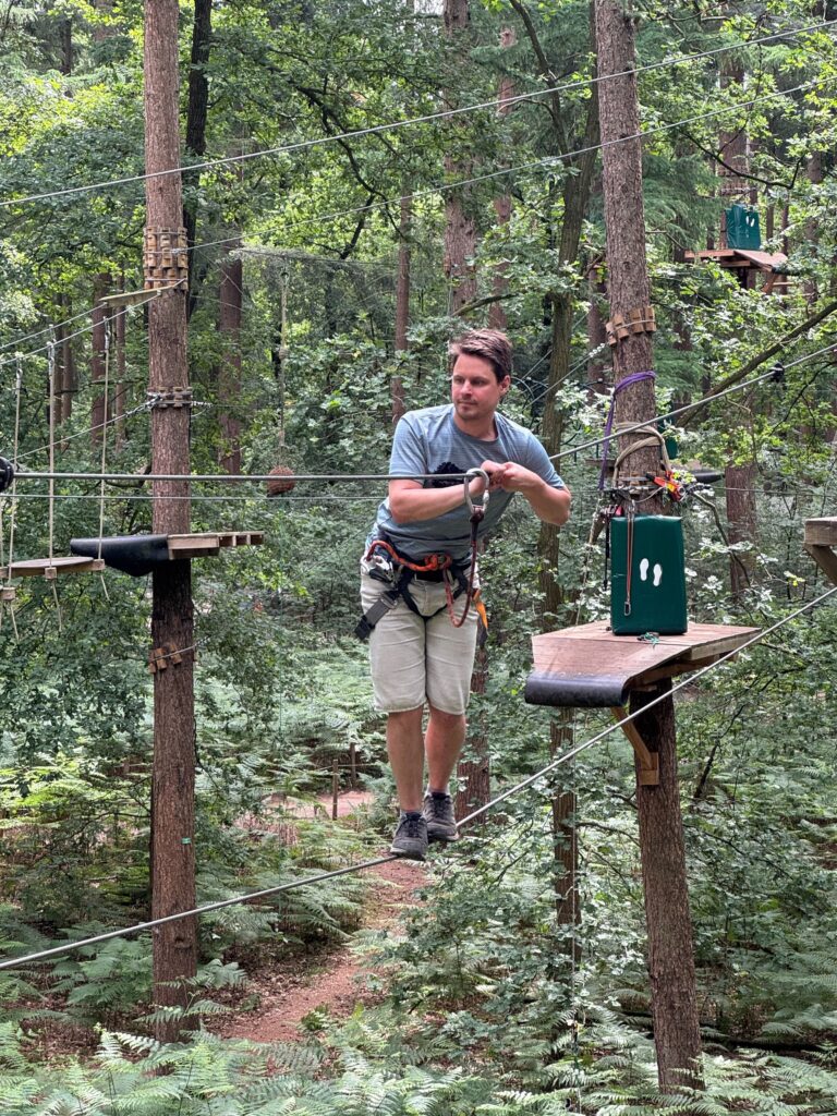 Persoon die geconcentreerd kijkt terwijl hij in klim-outfit over twee touwen balanceert. Bos op de achtergrond.