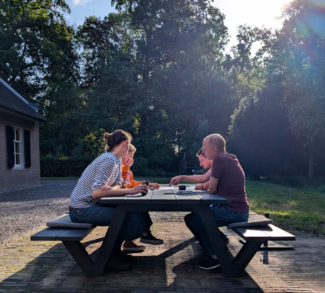 Vier mensen aan een picnictafel, met papieren in het midden. Zonnetje op de achtergrond.